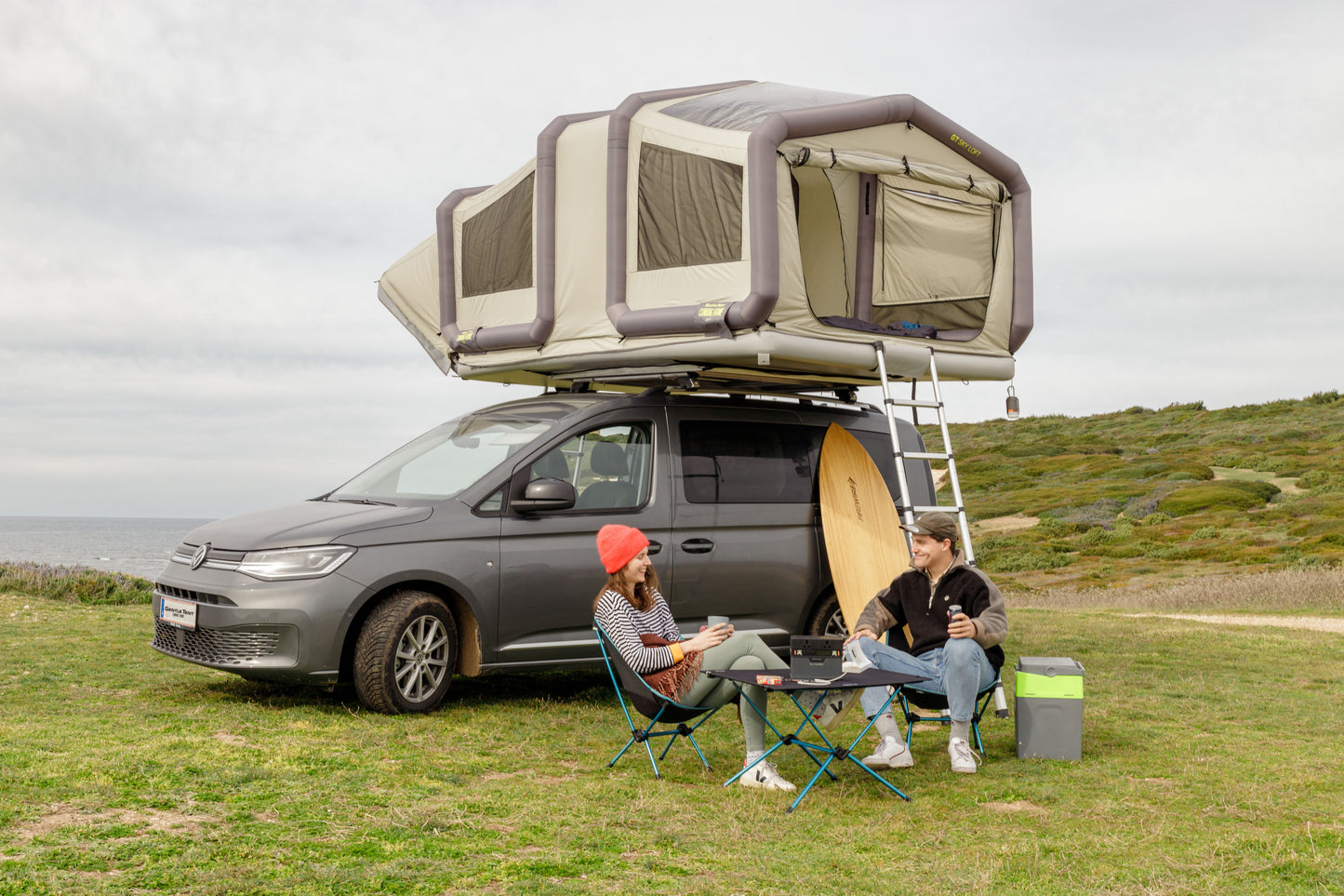 Olive SKY LOFT tent inflated and set up at a campsite by the ocean. Two campers sitting in front of the car enjoying their campaign trip with a surfboard in the background.