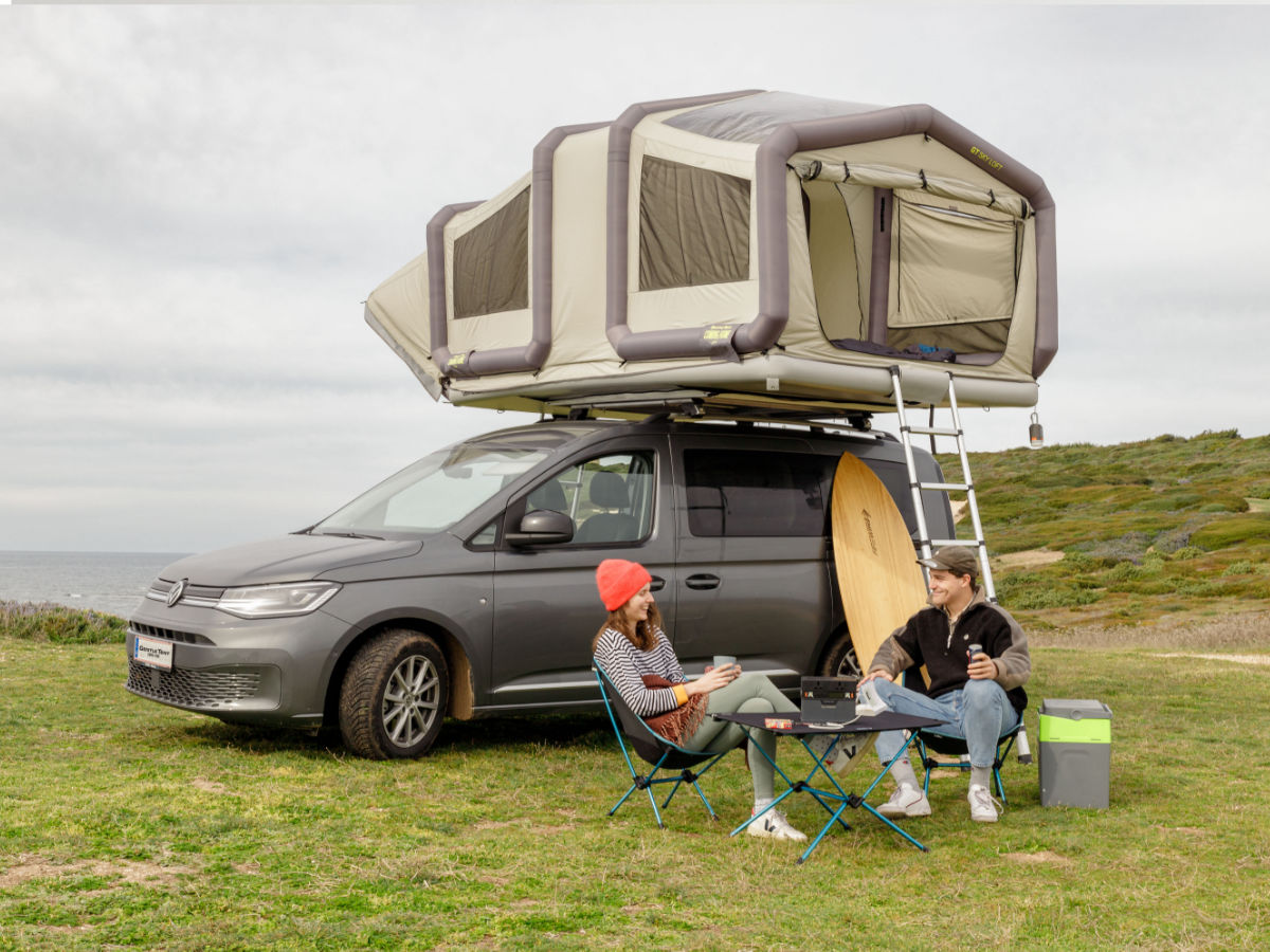 A couple sitting in camping chairs near the beack in front of their fully set up Sky Loft rooftop tent.