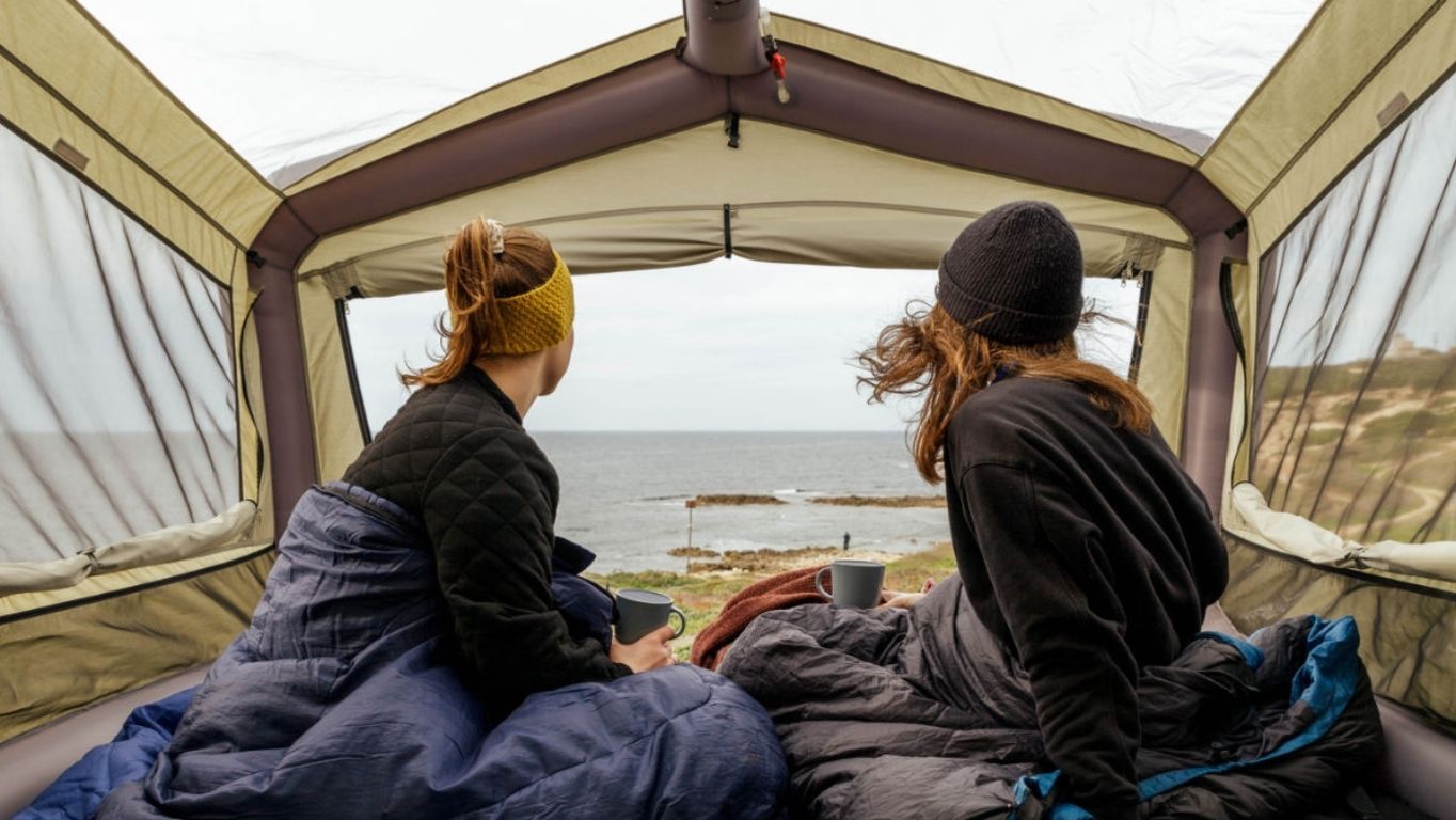 Two friends sitting in the Sky Loft rooftop tent enjoying an ocean view with their morning coffee.